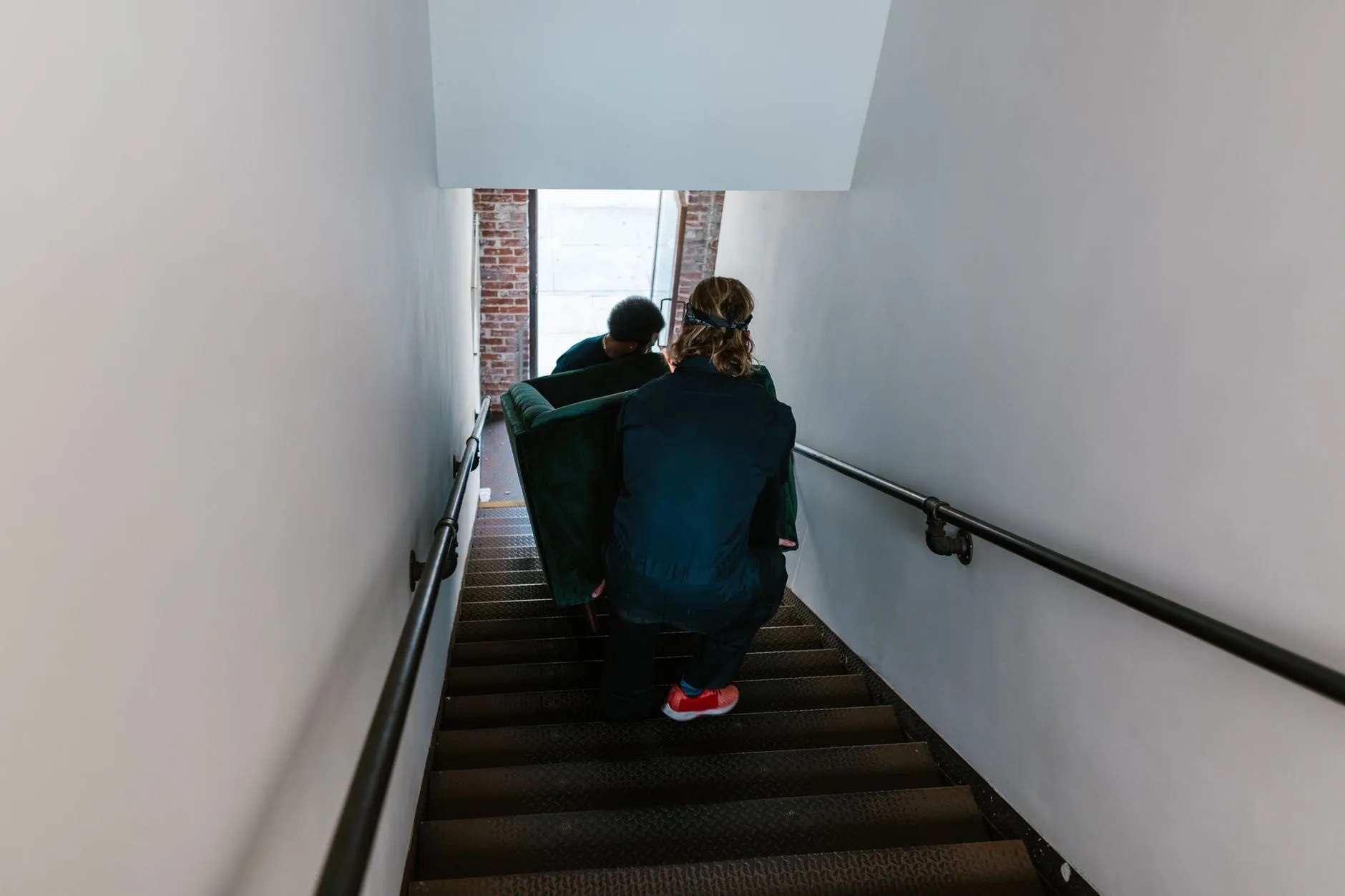 Two movers in navy work clothes carrying a green sofa down a narrow staircase, seen from behind